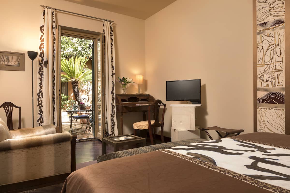 Habitación de hotel con cama, escritorio, silla y televisor. Se ve una ventana con cortinas, una lámpara y una planta.