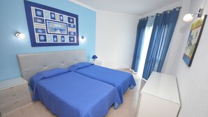 A bedroom with two beds, a nightstand, a dresser, a wall art piece, and a window with curtains. - Apartamentos Cravinho (Albufeira)