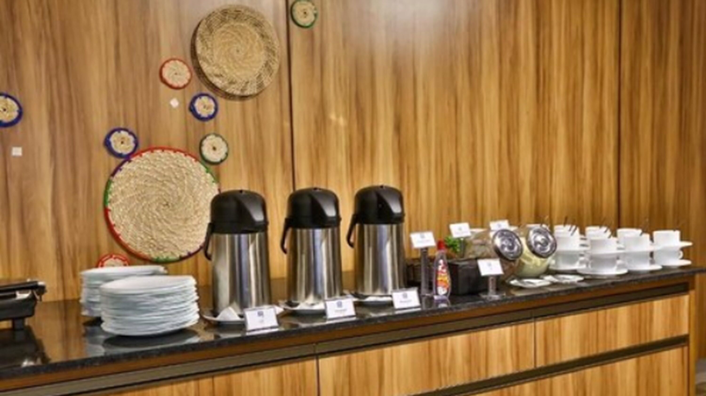 Una barra de desayuno con platos apilados, dos cafeteras de acero inoxidable y una variedad de tazas y platillos.