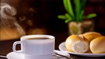 Eine weiße Kaffeetasse auf einem Untertasse, ein Teller mit Brötchen und eine grüne Pflanze im Hintergrund.