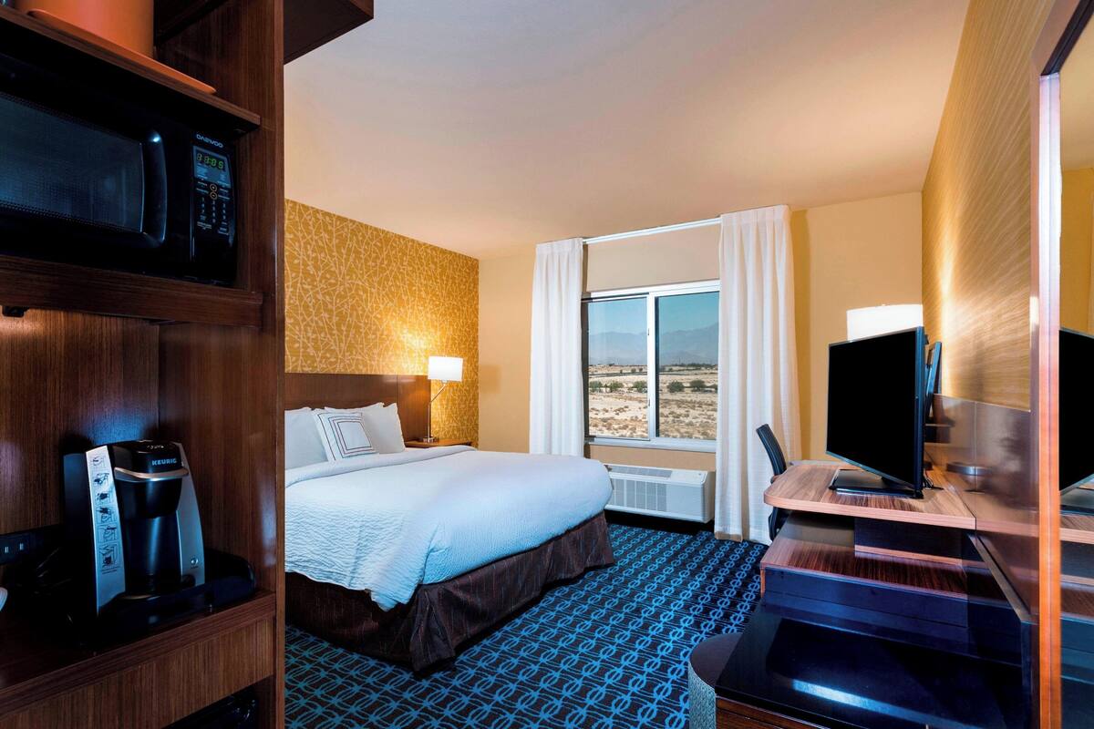 A hotel room with a bed, a desk with a computer, a coffee maker, a TV, and a window with a view of the desert.