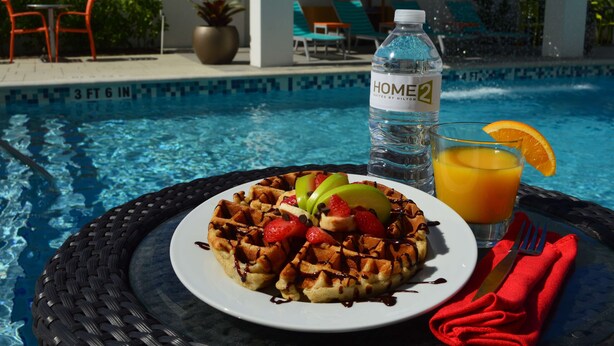 A dining table with a plate of waffles, a bottle of water, and a glass of orange juice by a pool. - Home2 Suites by Hilton Ft. Lauderdale Airport-Cruise Port (Dania Beach)