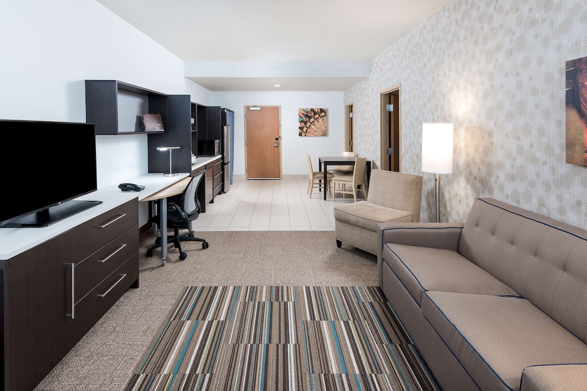 A modern hotel room with a flat-screen TV, a desk with a chair, a sofa, a patterned rug, and a dining area with a table and chairs.