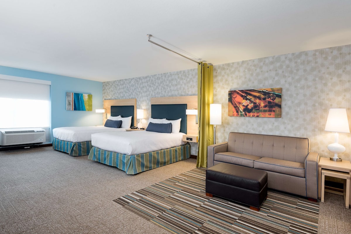A hotel room with two beds, a sofa, a coffee table, a TV, and a window with blinds.