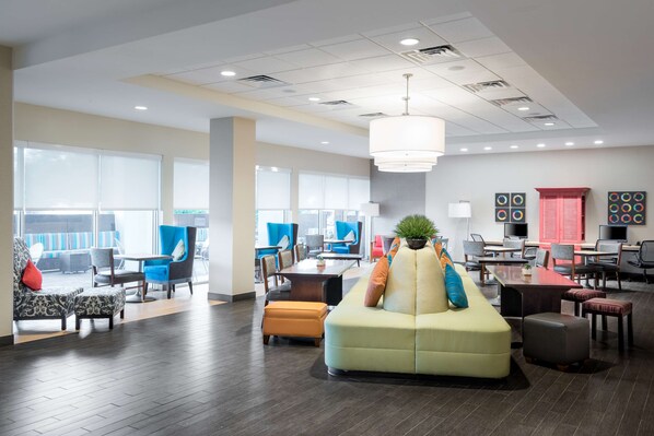 A spacious lobby with a central seating area, multiple tables, and chairs, and large windows providing natural light.