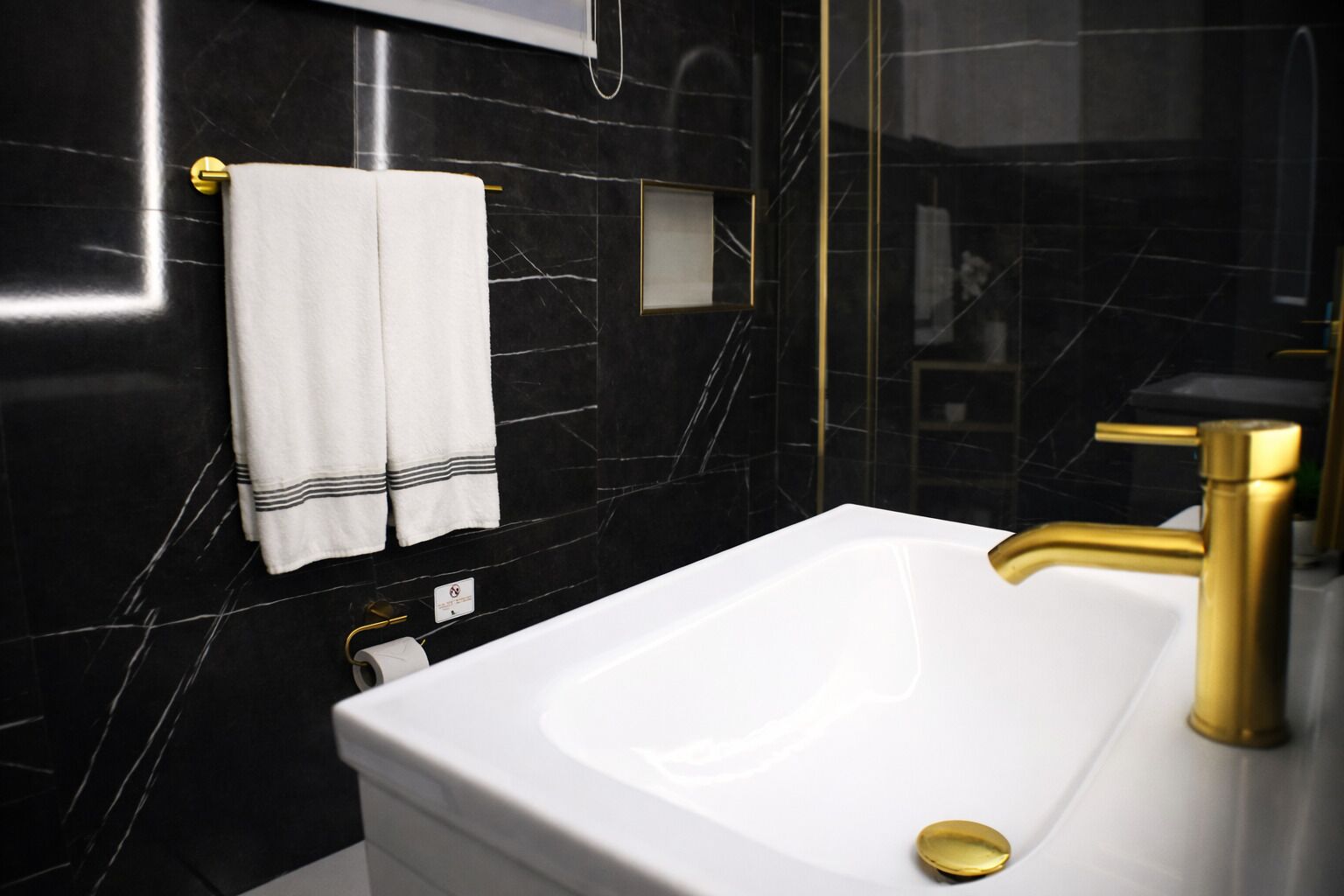 Un baño moderno con un lavamanos blanco, grifo dorado y paredes de mármol negro.