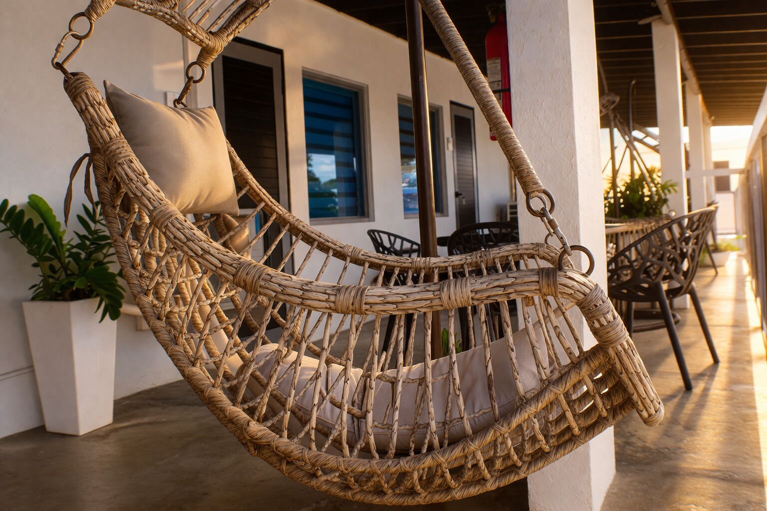 A wicker hammock chair with a cushion, positioned on a concrete floor, with a building and outdoor furniture in the background.