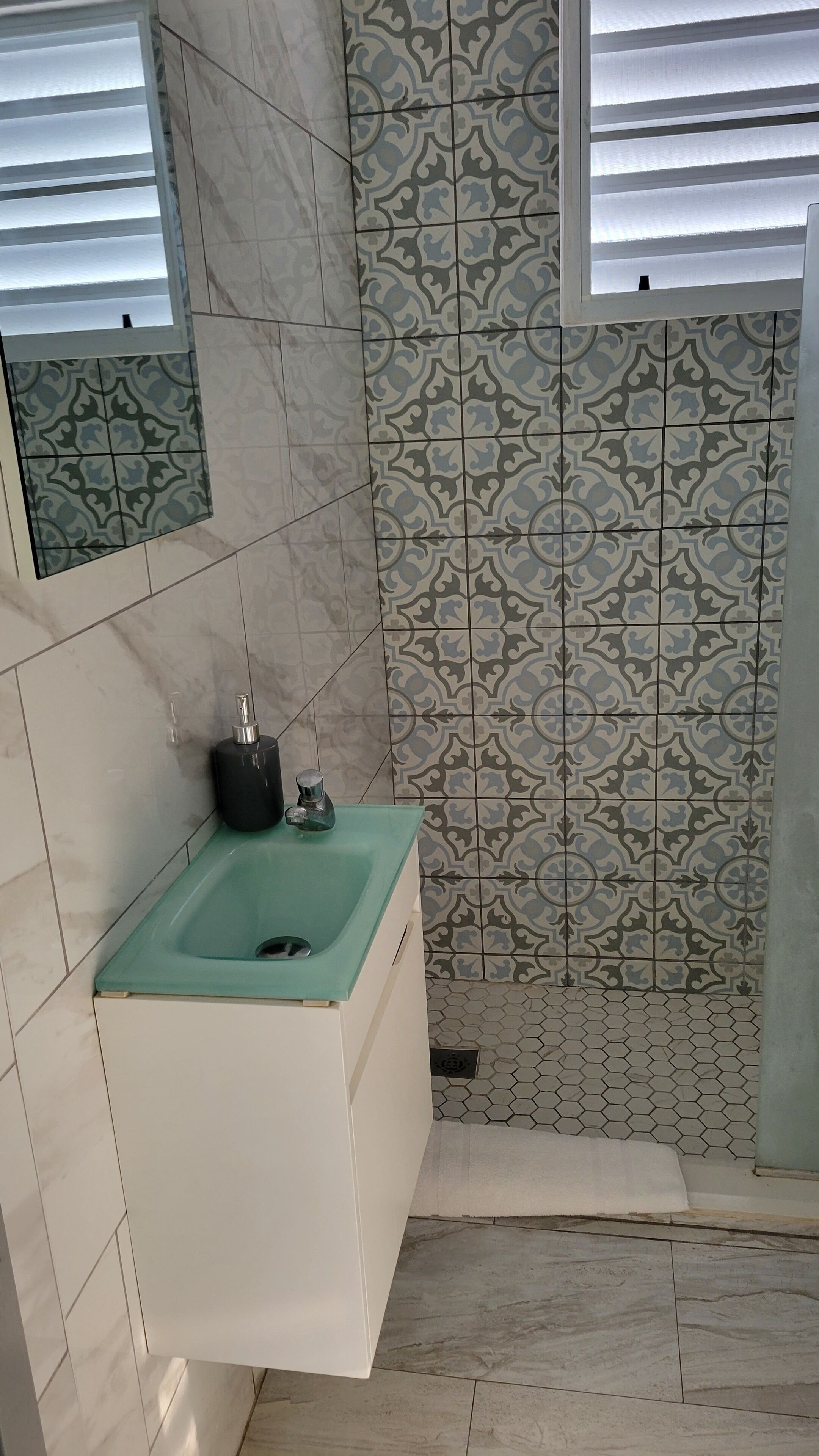 Une salle de bain avec un lavabo vert, des carreaux à motifs et une fenêtre avec des stores.