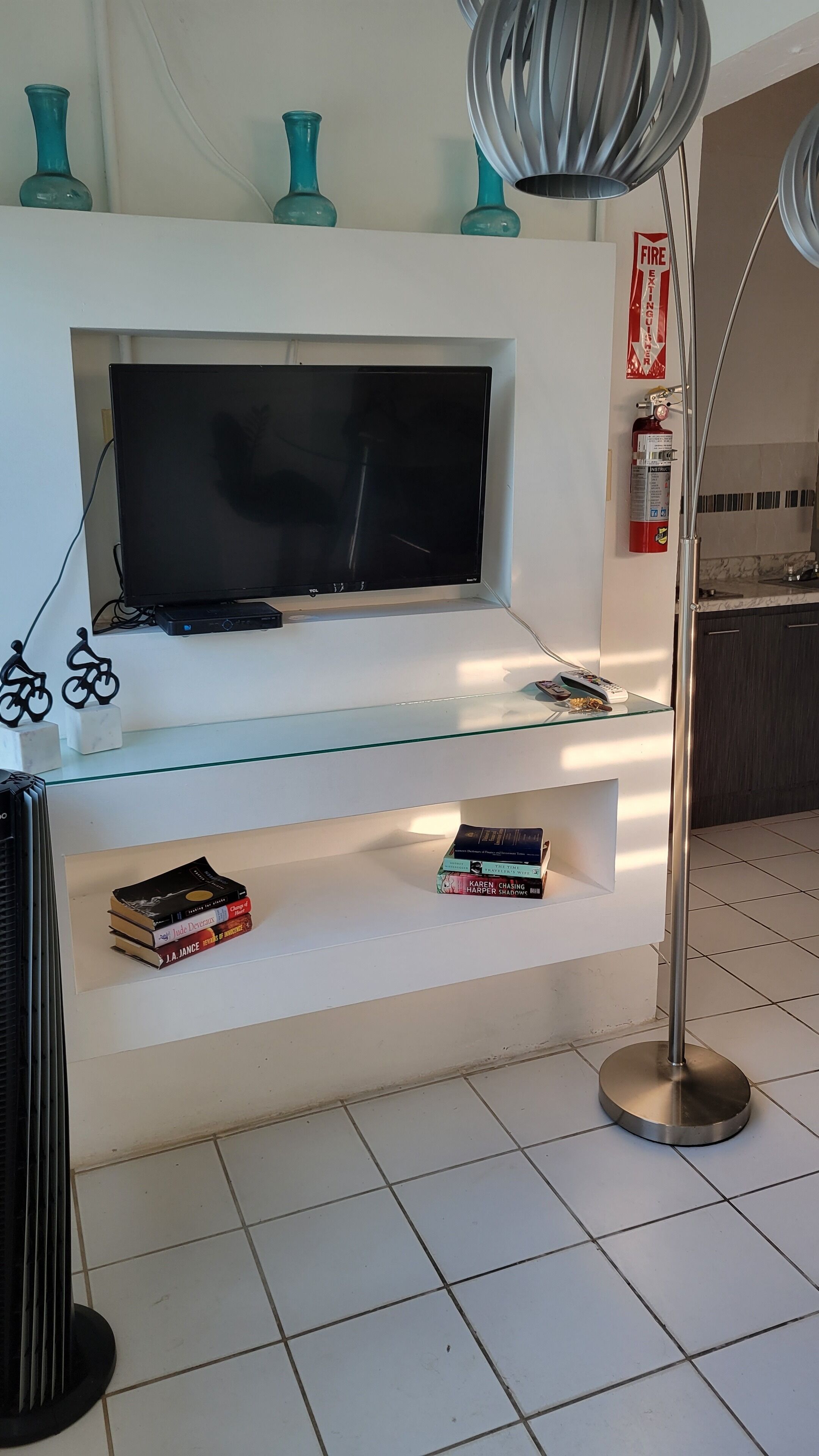 A living room with a flat-screen TV mounted on a white wall, a glass shelf with books, a floor lamp, and a fire extinguisher.