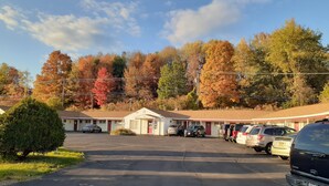 A motel with a parking lot, several cars parked in front, and autumn-colored trees in the background. - Cortland Motel (Cortland)