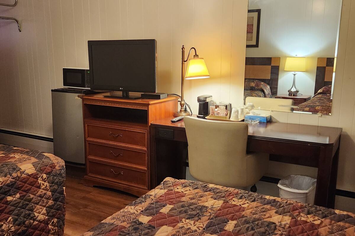 A hotel room with two beds, a desk with a computer, a lamp, a coffee maker, and a trash bin.