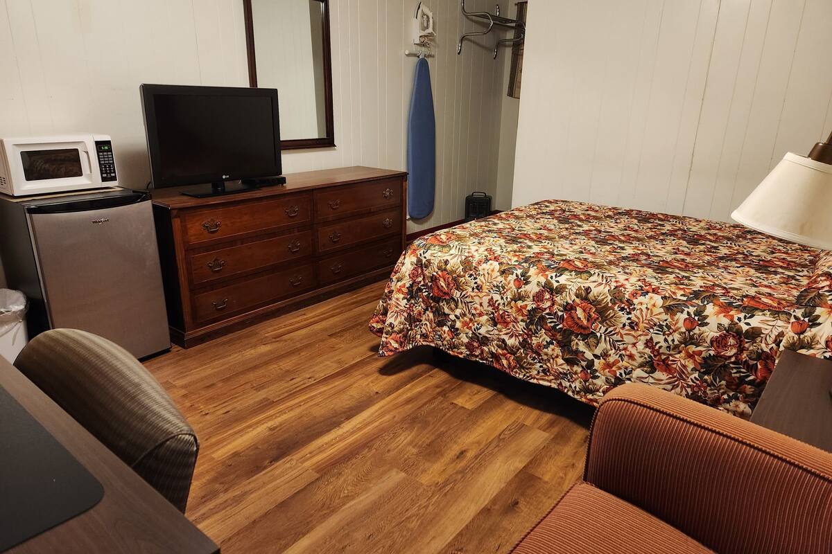 A bedroom with a wooden floor, a bed with a floral bedspread, a dresser, a television, a microwave, and a small refrigerator.