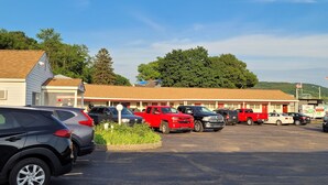 A parking lot with several cars, a building with a red roof, and trees in the background. - Cortland Motel (Cortland)