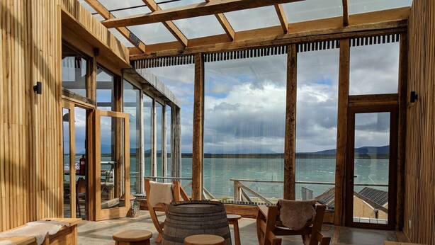 A wooden deck with a glass enclosure, wooden stools, a barrel, and a rocking chair, overlooking a body of water. - Hotel Simple Patagonia (Natales)