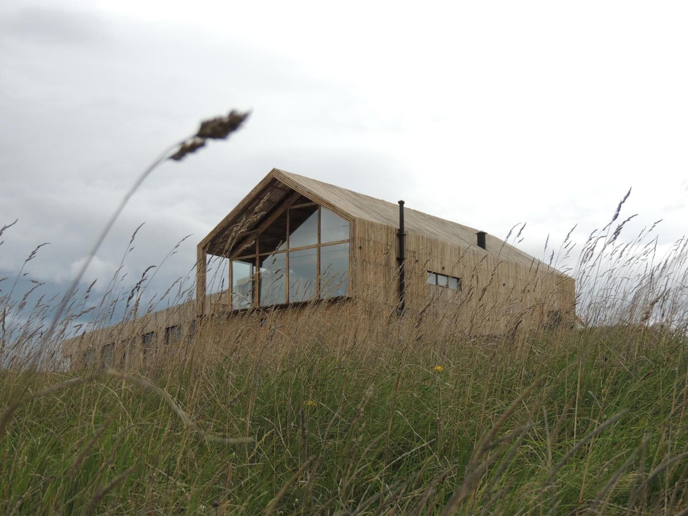 A modern, minimalist building with large windows and a sloped roof, surrounded by tall grass and a cloudy sky.