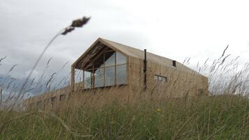 A modern, minimalist building with large windows and a sloped roof, surrounded by tall grass and a cloudy sky.