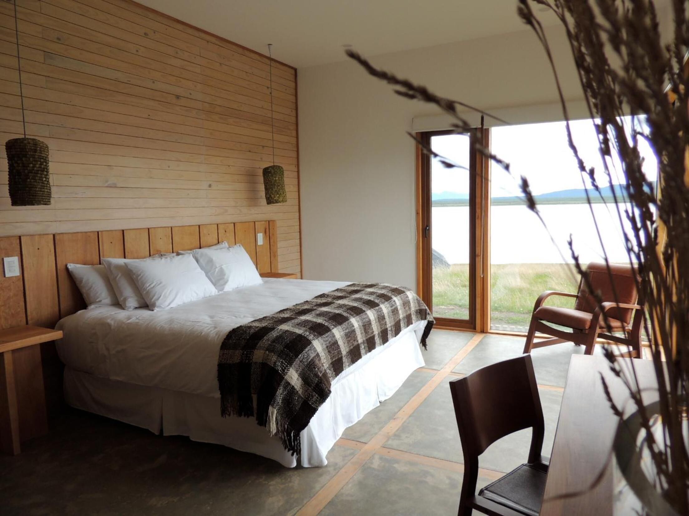 Un dormitorio con cabecera de madera, una cama grande con ropa blanca, un plaid de cuadros y vistas a un lago a través de una puerta corrediza.