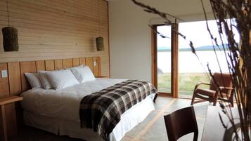Un dormitorio con cabecera de madera, una cama grande con ropa blanca, un plaid de cuadros y vistas a un lago a través de una puerta corrediza.