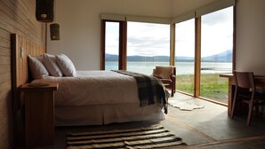 A bedroom with a large bed, a wooden headboard, a bedside table, a chair, a desk, and a view of a lake through large windows. - Hotel Simple Patagonia (Natales)