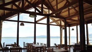 A spacious dining area with wooden tables and chairs, large windows offering a view of a lake and mountains. - Hotel Simple Patagonia (Natales)