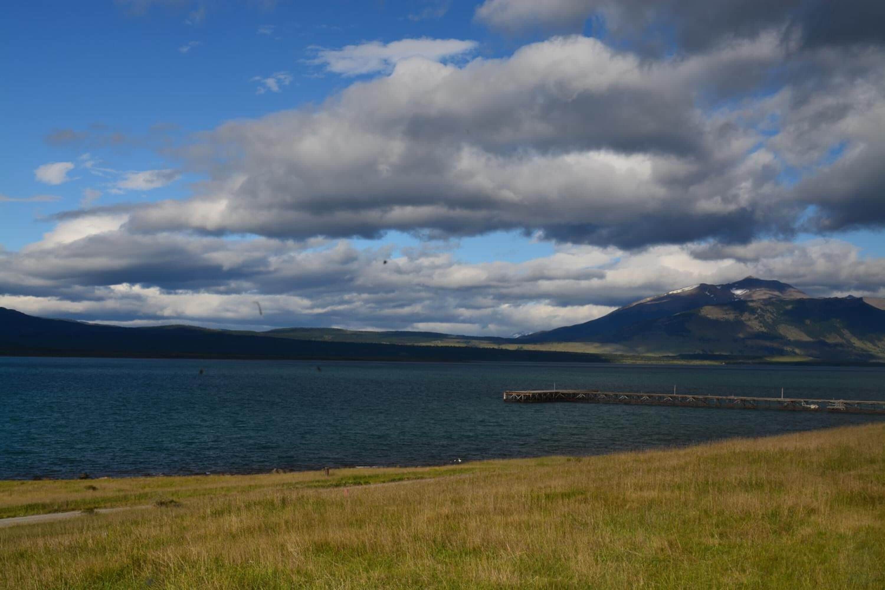 A lake with a dock extending into the water, surrounded by grassy areas and mountains in the background.