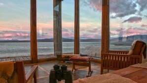 A cozy room with a large window overlooking a body of water and mountains. There are wooden chairs and a plush sofa. - Hotel Simple Patagonia (Natales)