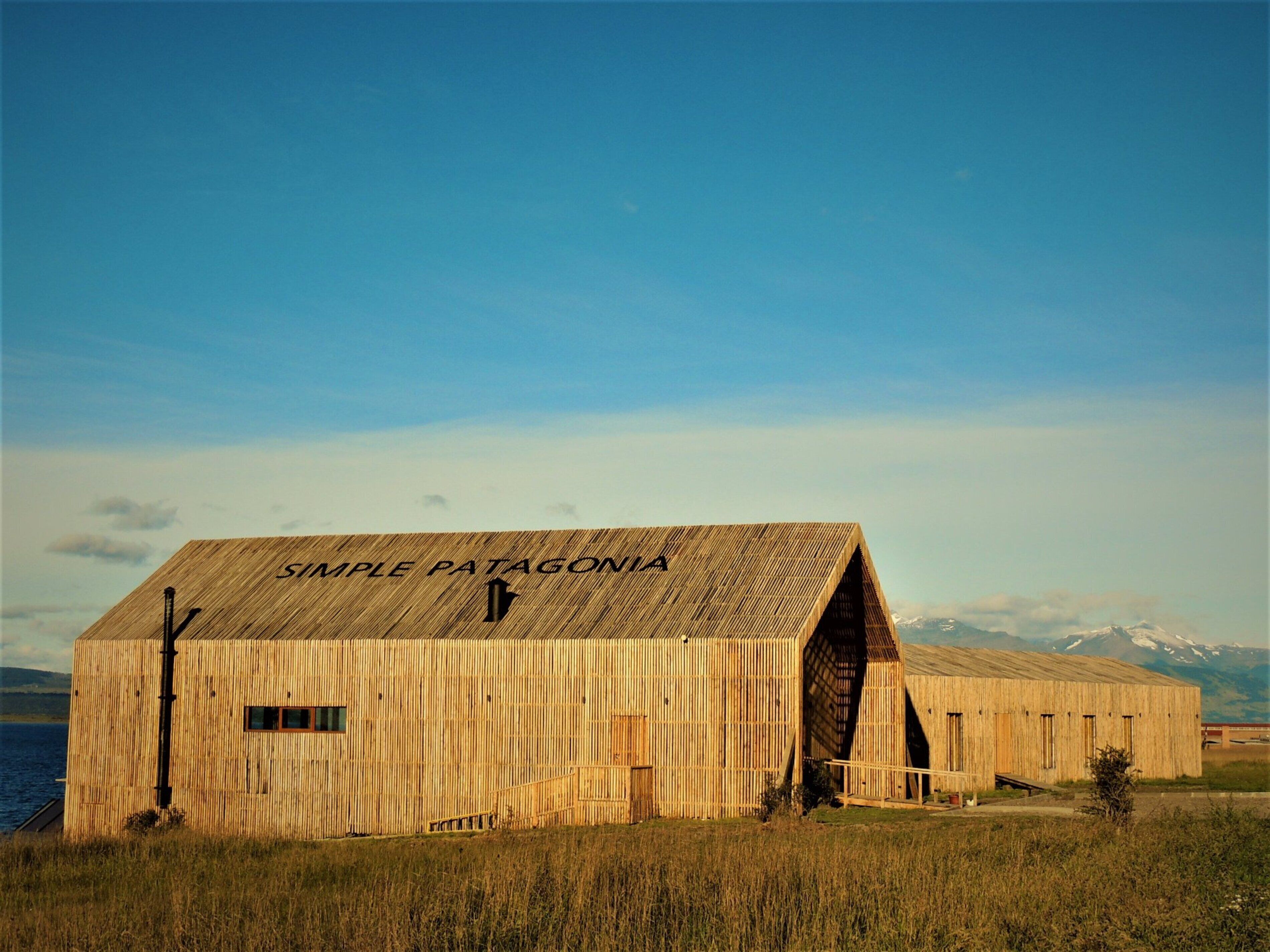 A rustic wooden building with the text "SIMPLE PATAGONIA" on its facade, located near a body of water with mountains in the background.