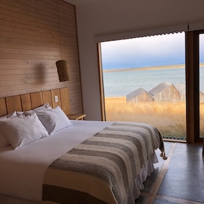 A bedroom with a large bed, wooden headboard, and a view of the sea through a large window. - Hotel Simple Patagonia (Natales)