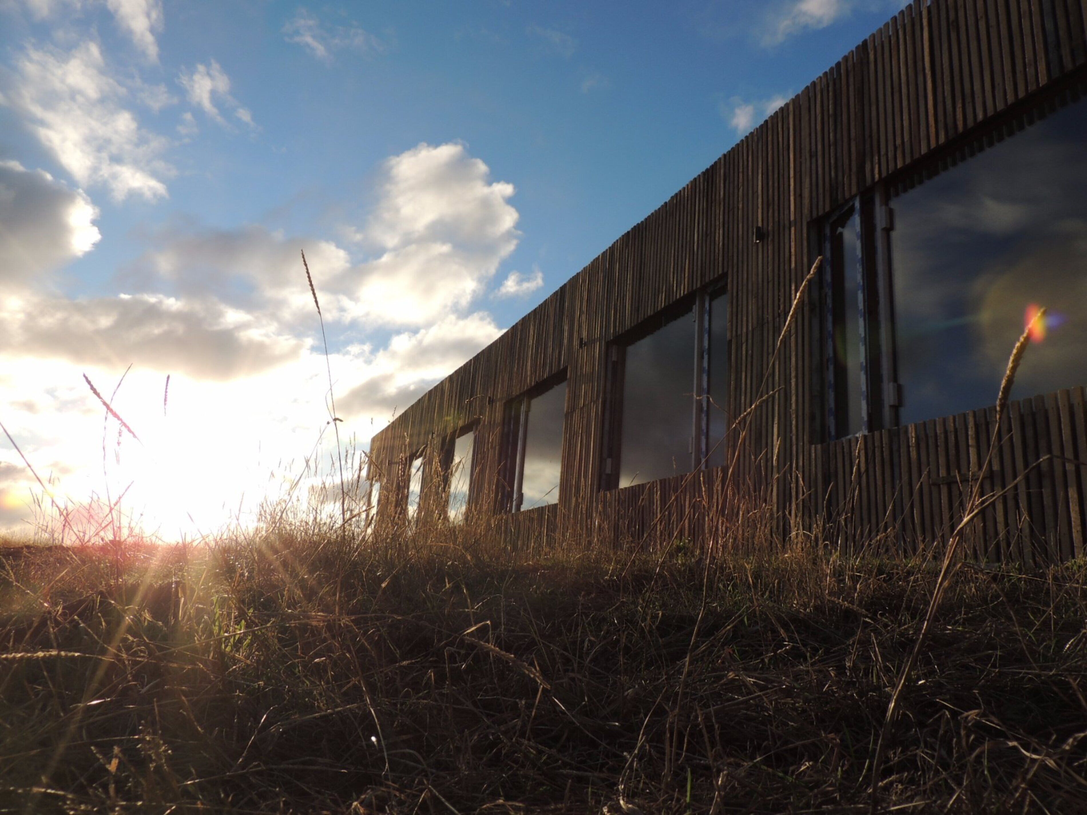 A modern building with a wooden facade and large windows, set against a backdrop of a clear sky and sunlight.