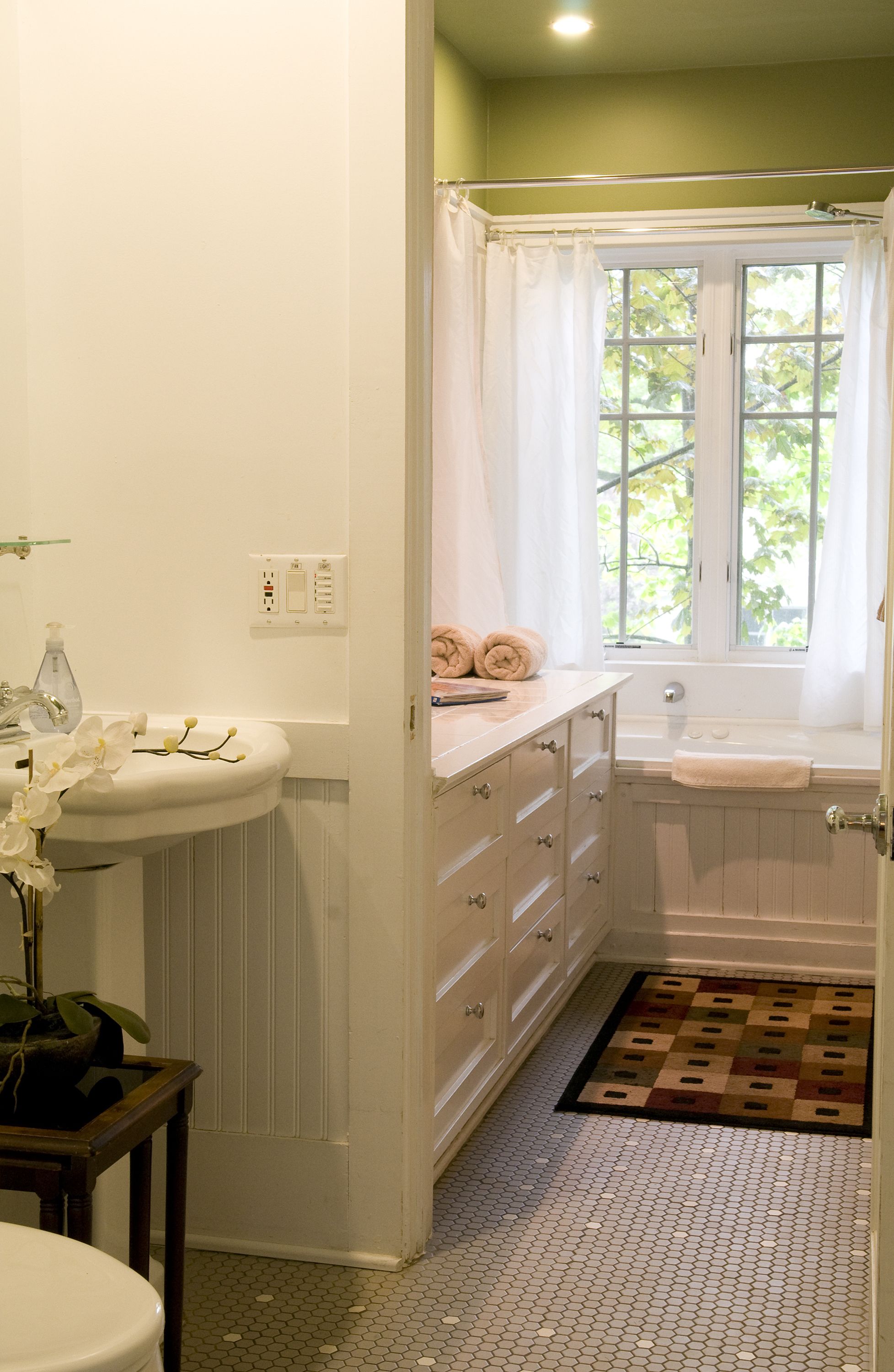 A bathroom with a white sink, a bathtub, a window with a view of trees, and a patterned rug on the floor.