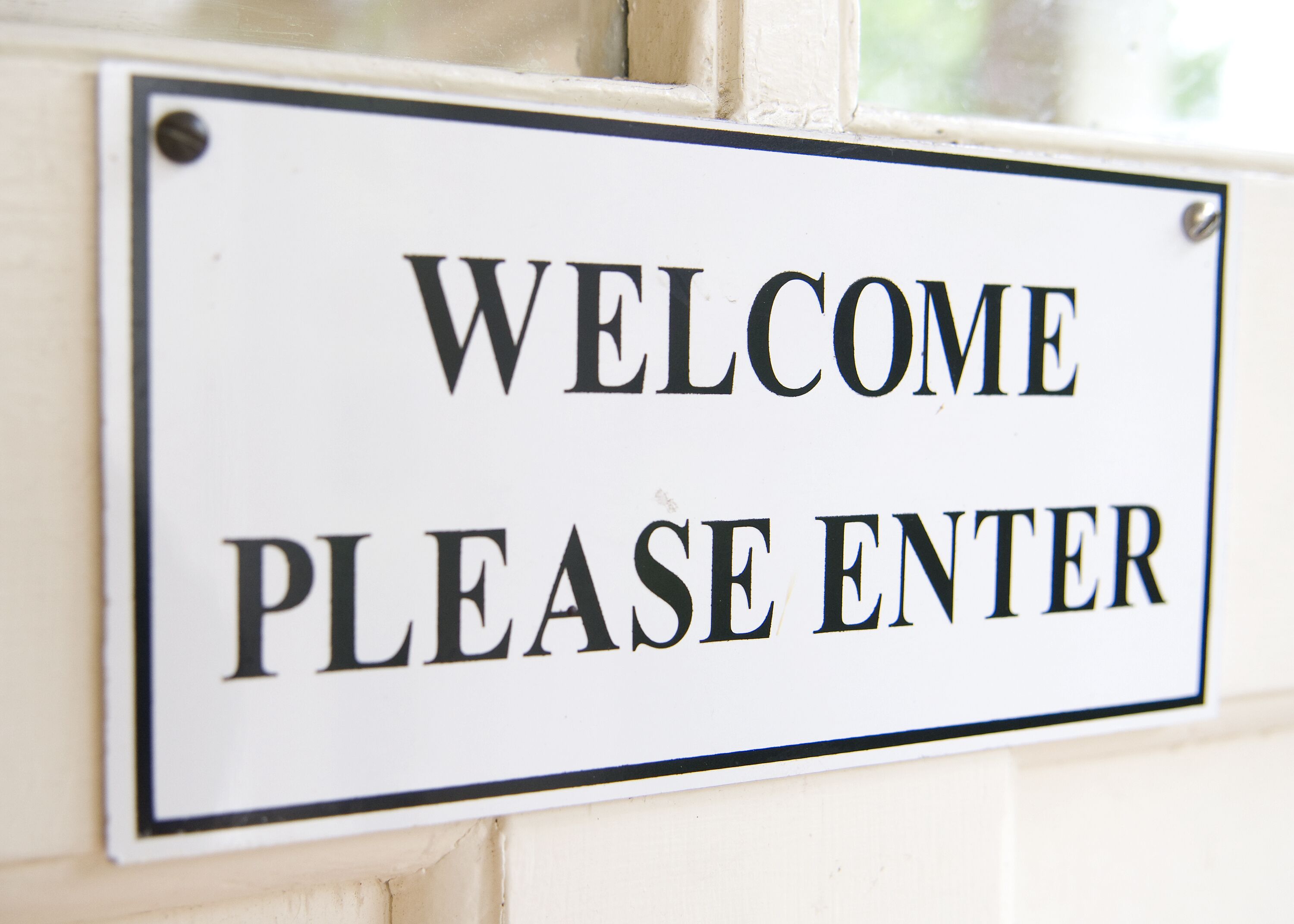 A welcome sign with the text "WELCOME PLEASE ENTER" on a white background with a black border, affixed to a wall.