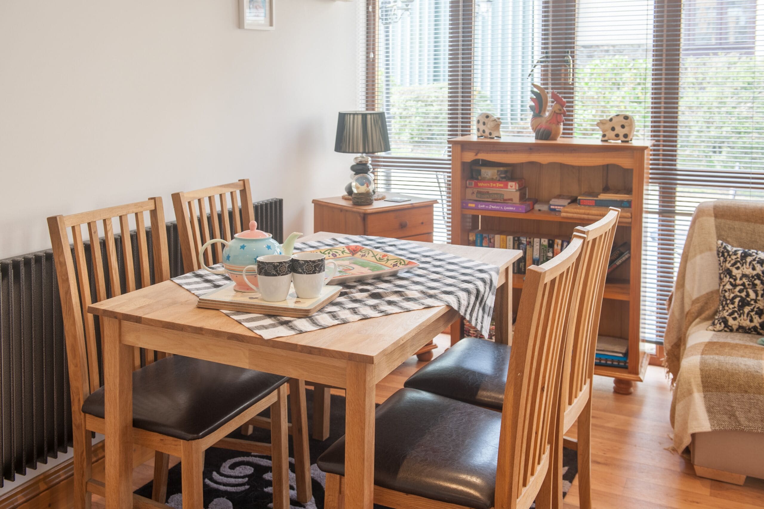 Un coin repas accueillant avec une table en bois, des chaises noires à coussins et une nappe à carreaux. On aperçoit, à l’arrière-plan, une bibliothèque remplie de livres et d’objets décoratifs.