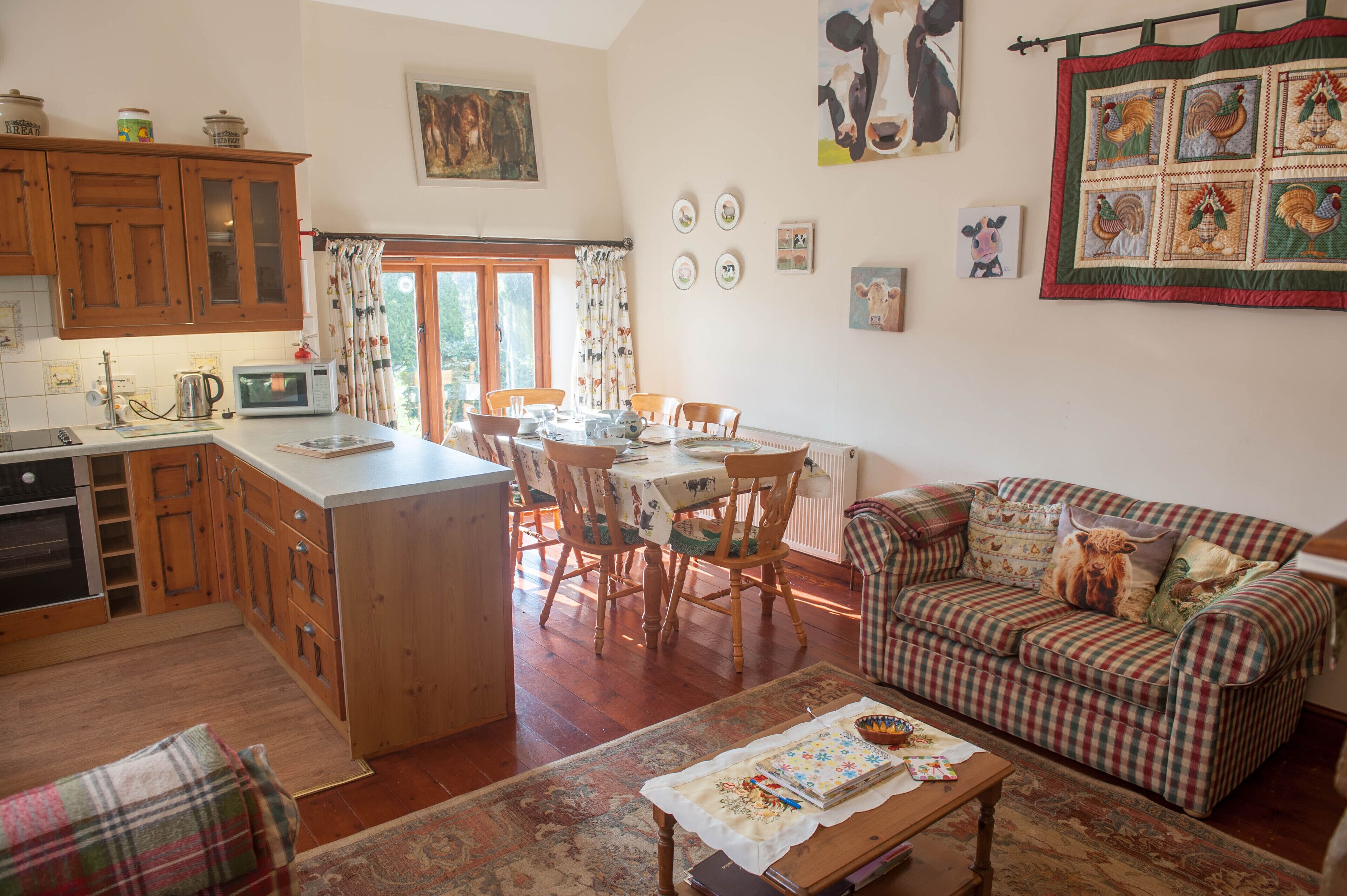 A cozy living room with a checkered sofa, a dining table with chairs, and a kitchen area with wooden cabinets and a microwave.