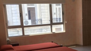 Una habitación con dos camas, una ventana con vistas a edificios, un ventilador de techo y un equipo de aire acondicionado.