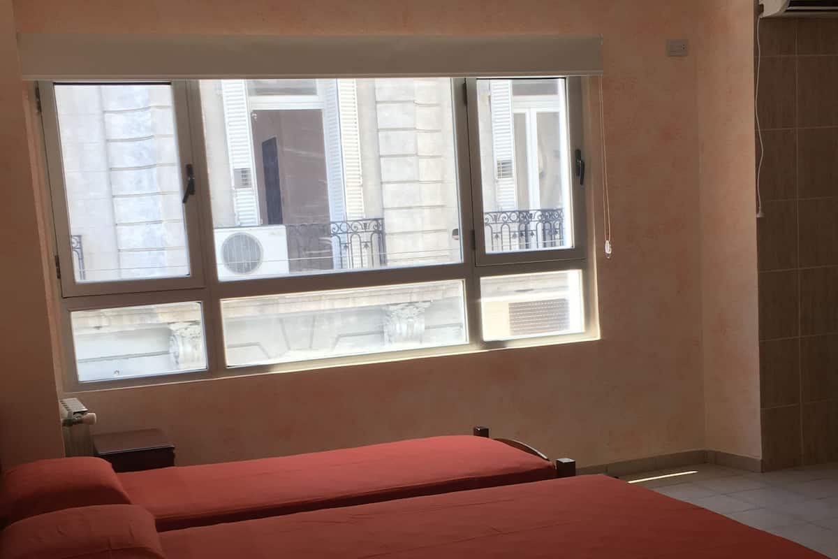 A room with two beds, a window with a view of buildings, a ceiling fan, and an air conditioning unit.