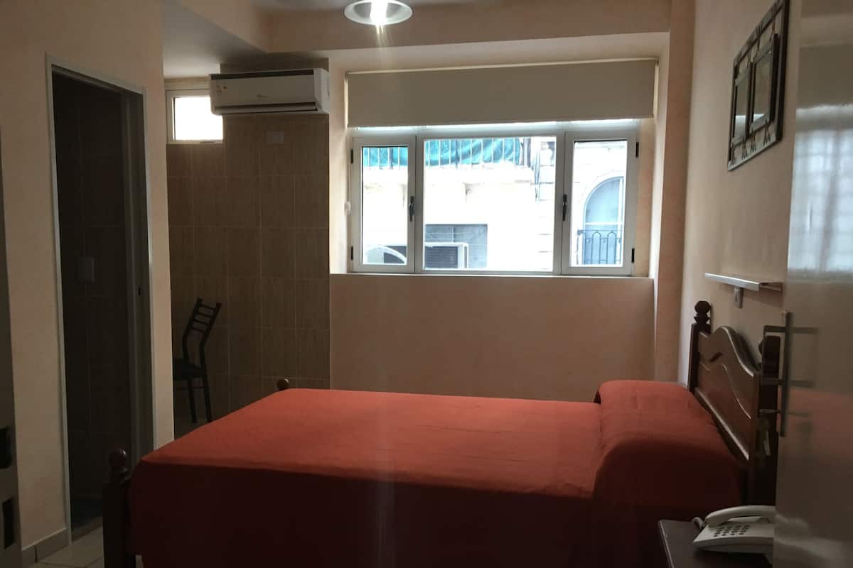 A bedroom with a bed, a window, a chair, and a bedside table with a phone.