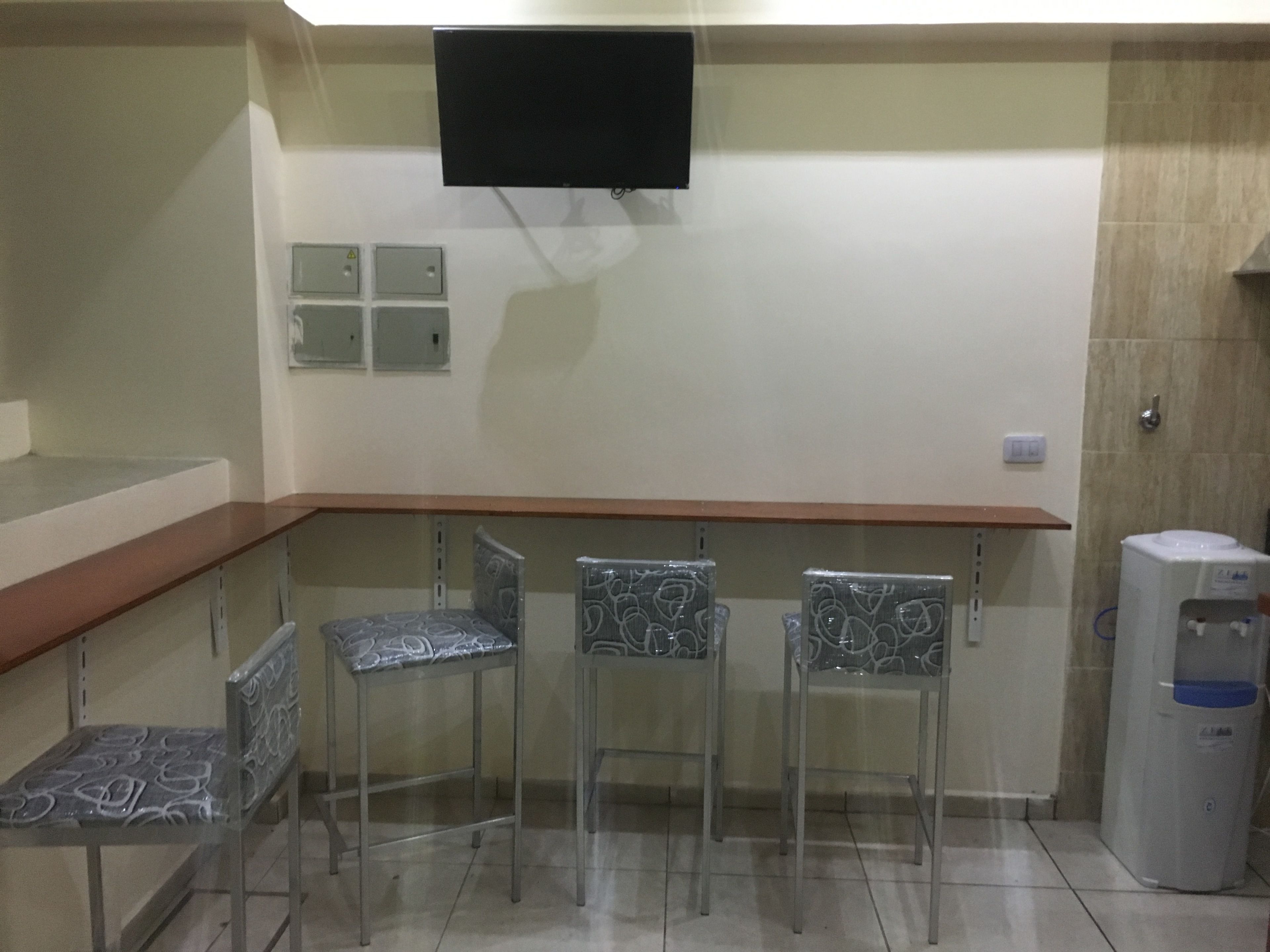 A small bar area with a flat-screen TV mounted on the wall, a wooden counter, and three chairs with patterned upholstery.