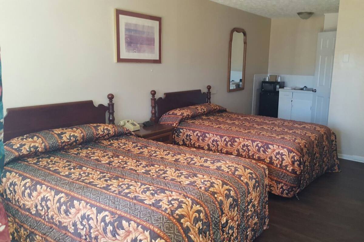 A hotel room with two beds, a desk, a chair, a mirror, a framed picture on the wall, and a kitchenette area.
