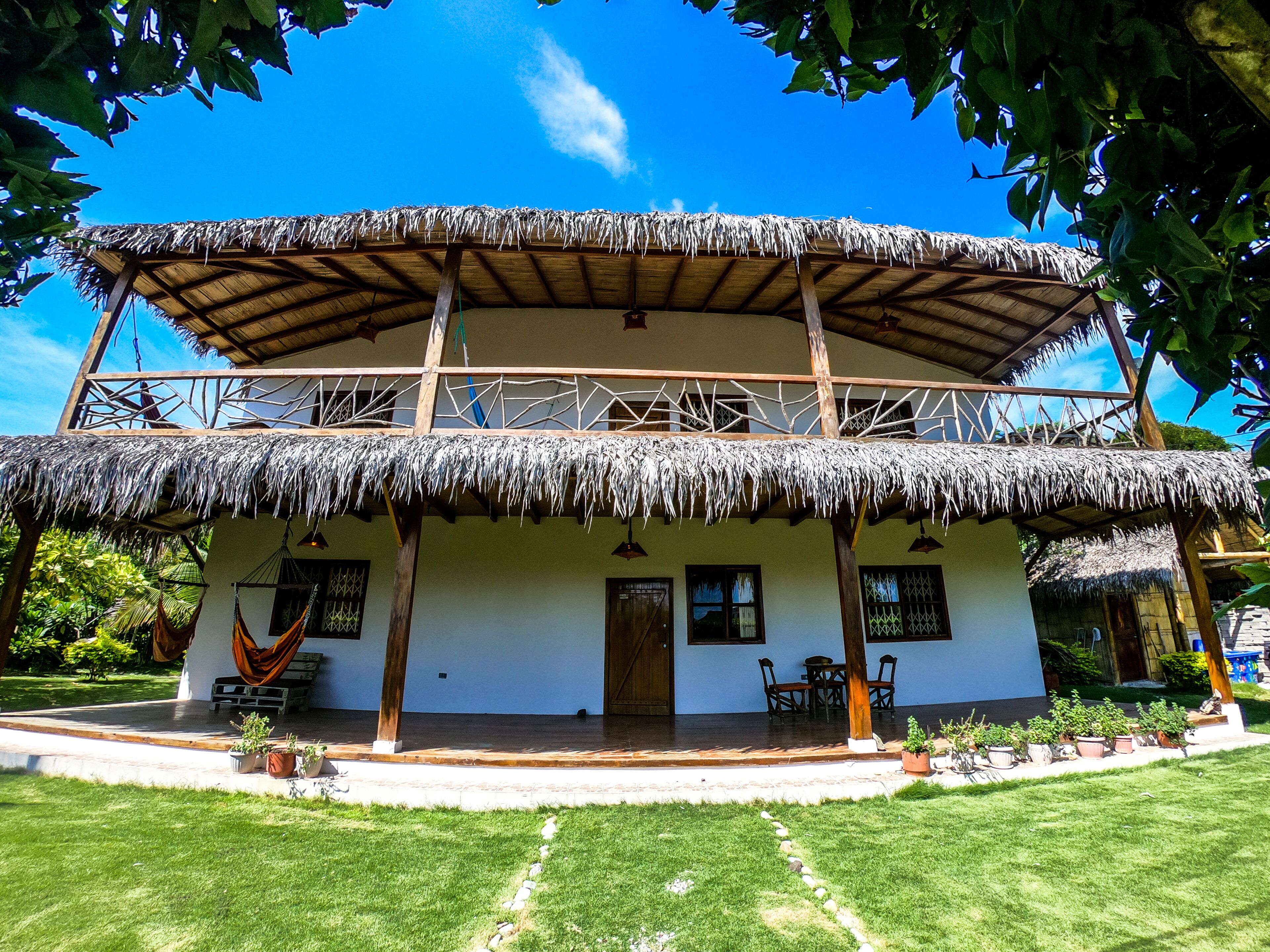 A two-story building with a thatched roof, wooden balconies, and a garden area with potted plants.