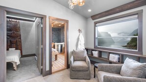 Een moderne kamer met een groot raam, een houten deur naar een badkamer en een zithoek met uitzicht op een bergachtig landschap.