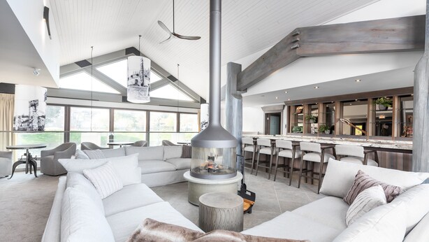 Een ruime woonkamer met een grote witte bank, een ronde salontafel en een moderne haard. Er is een keuken met barstoelen en een groot raam dat voor natuurlijk licht zorgt.