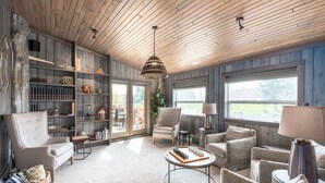 Een ruime woonkamer met een hoge plafond, houten balken en grote ramen. Er zijn meerdere zithoeken met banken en fauteuils, een salontafel en een boekenkast vol boeken en decoratieve objecten.