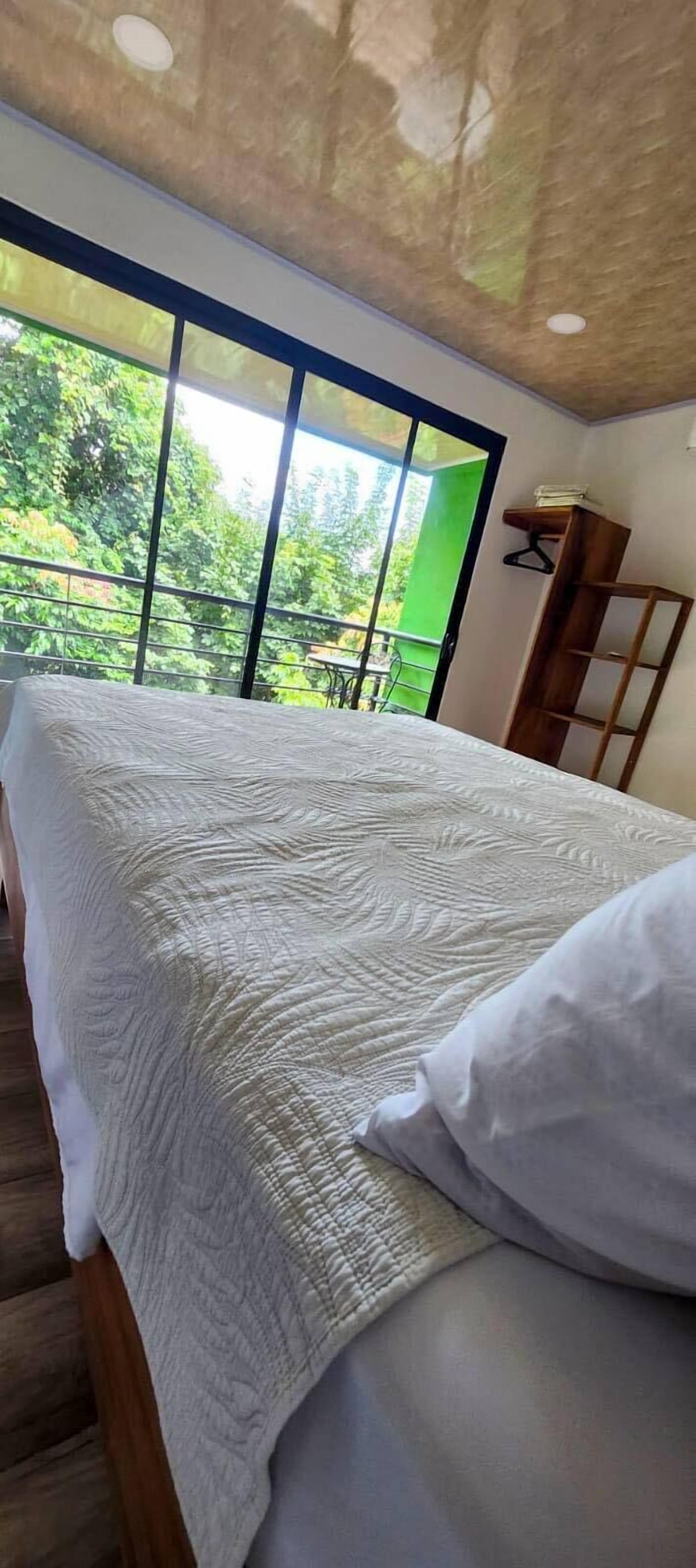 A bed with a white quilt and pillow, a wooden headboard, and a view of greenery through large windows.