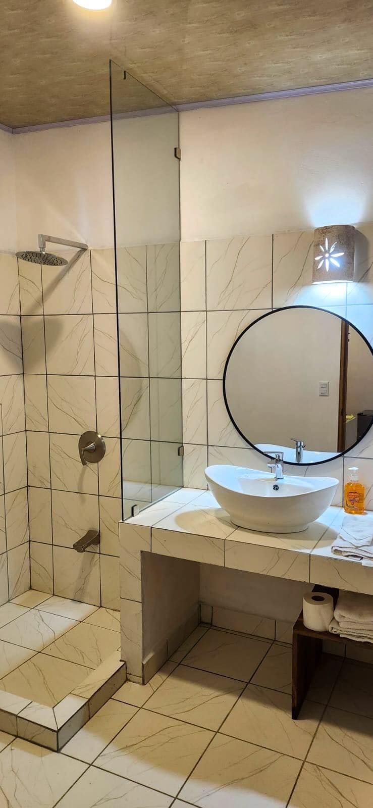 A modern bathroom with a glass-enclosed shower, a round mirror, and a white sink.
