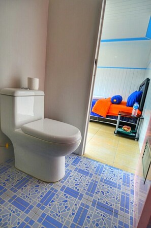 A bathroom with a toilet, a blue and white tiled floor, and a bed with an orange bedspread in the adjacent room.