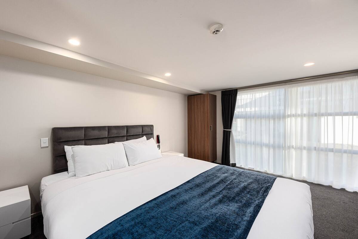 A modern hotel room with a large bed, a bedside table, a wardrobe, and a window with sheer curtains.