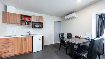 Cozinha compacta com micro-ondas, chaleira e frigorífico, uma mesa de jantar com cadeiras e uma janela com cortinas.