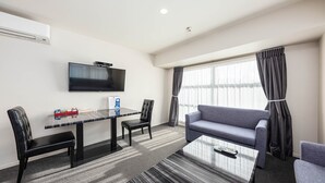 Ein modernes Hotelzimmer mit einem an der Wand befestigten Flachbildfernseher, einem Esstisch mit Stühlen, einem Sofa und einer Fernbedienung auf einem Glastisch.