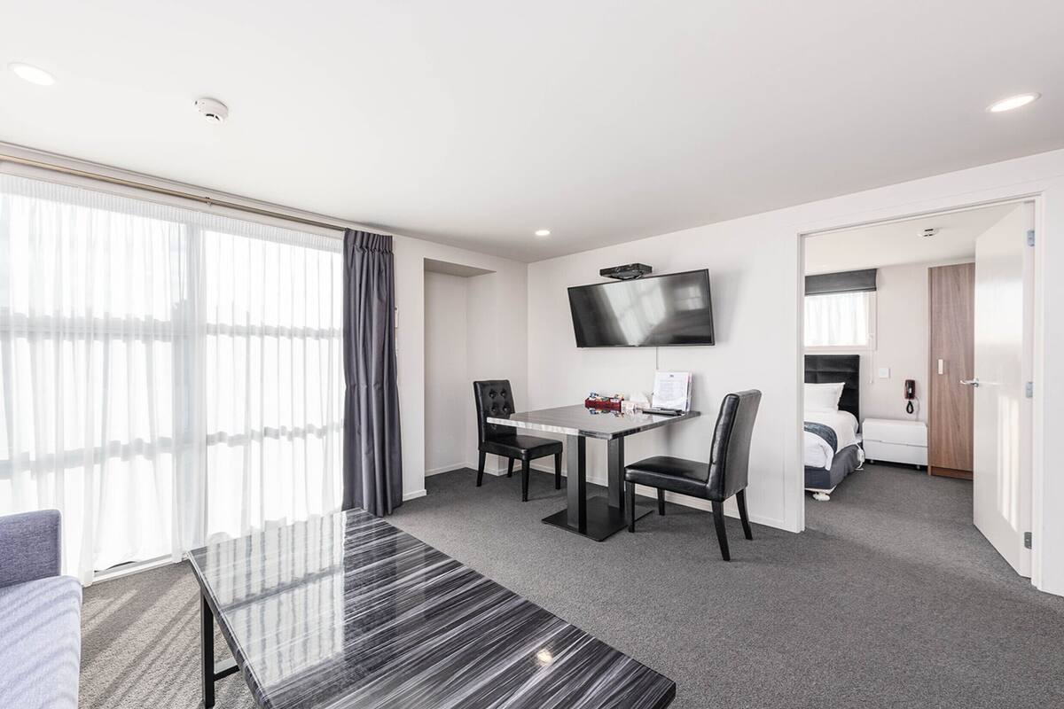 A hotel room with a dining area, a TV mounted on the wall, a sofa, and a bed.