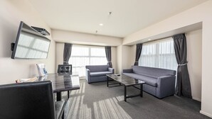 Ein modernes Hotelzimmer mit einem an der Wand befestigten Flachbildfernseher, einer Couch, zwei Sesseln, einem Couchtisch und einem kleinen Schreibtisch mit Stuhl.